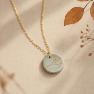 May include: A gold chain necklace featuring a circular, light sage green ceramic pendant. The pendant is decorated with a thin, branching gold-colored line pattern that resembles a crack or a twig across its smooth, matte surface.