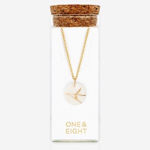 May include: A gold chain necklace with a white circular pendant featuring a delicate gold leaf design. The necklace is displayed in a clear glass jar with a cork top. The label on the jar reads "ONE & EIGHT".