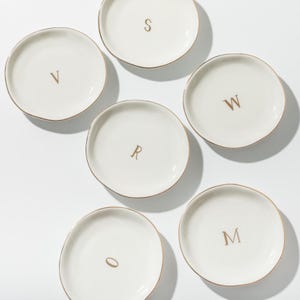 May include: Six small, round, white ceramic plates with gold rims, each bearing a single gold letter: V, S, W, R, O, and M. The plates are arranged on a white surface, offering a minimalist aesthetic.