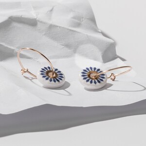 May include: A pair of gold hoop earrings with white ceramic charms. The charms feature a blue and gold geometric pattern.