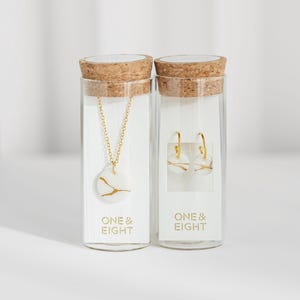May include: Two glass vials with cork stoppers, each containing jewelry. One vial holds a gold chain necklace with a white pendant and gold design. The other vial contains gold hoop earrings with a white and gold design. The text "ONE & EIGHT" is printed on the front of each vial.