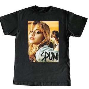 Puede incluir: Camiseta negra con un estampado gráfico de una mujer con el pelo largo y rubio y un hombre detrás de ella. El texto "SPUN" está impreso en la camiseta.