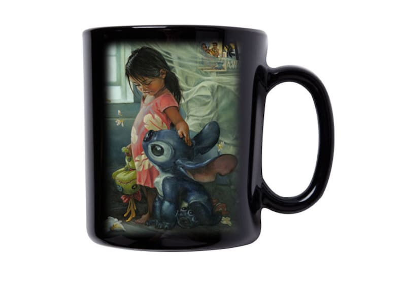 May include: Black ceramic mug with a colorful illustration of a young girl and a blue alien character. The girl is wearing a pink dress with floral patterns and the alien has large eyes and blue fur. The background is a soft, blurry image of a bedroom.