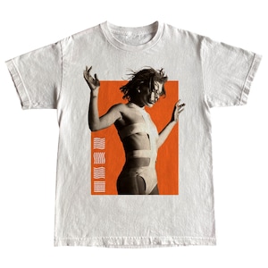 May include: White t-shirt with a black and white image of a person in a white outfit, set against an orange background. The design includes vertical lines on the left side of the orange rectangle. A stylish graphic tee.
