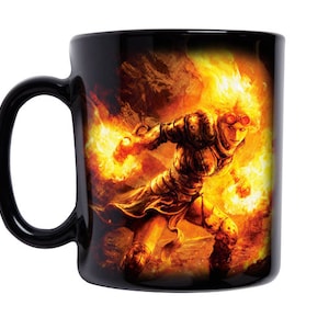 Puede incluir: Taza de cerámica negra con una imagen de fuego naranja y amarillo de una persona con llamas en el pelo y un orbe naranja brillante en su mano.