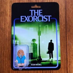May include: A vintage-style action figure card featuring Regan MacNeil from the movie "The Exorcist". The card has a blue and white border with a green and white background image. The figure is a small, blue-clad girl with brown hair.