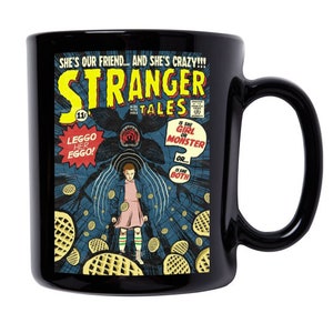 May include: Black ceramic mug with a Stranger Things comic book cover design. The cover features a girl with a pink dress and a monster in the background. The text on the cover reads "Stranger Tales" and "Is she girl or monster? Or... Is she both?"