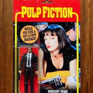 May include: A retro-style action figure packaging featuring Vincent Vega from the film Pulp Fiction. The packaging is red with yellow text and features a photo of Uma Thurman as Mia Wallace. The text on the packaging reads "Pulp Fiction" and "Would you give a guy a foot massage?"  The action figure is dressed in a black suit and tie and is holding a gun.