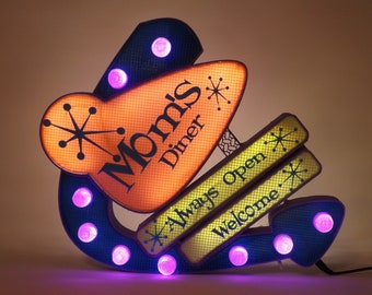 May include: A decorative wall sign shaped like a boomerang with blue, orange, and yellow panels. It features the text "Mom's Diner," "Always Open," and "Welcome" in black lettering, accented by starburst graphics and illuminated by purple bulbs.