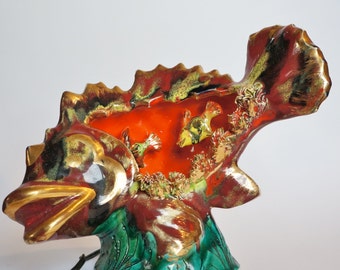 Può includere: Una lampada a forma di pesce in ceramica con una glassa rossa, arancione e verde. Il pesce ha dettagli dorati ed è decorato con pesci più piccoli e coralli. La lampada ha un cavo e un interruttore.