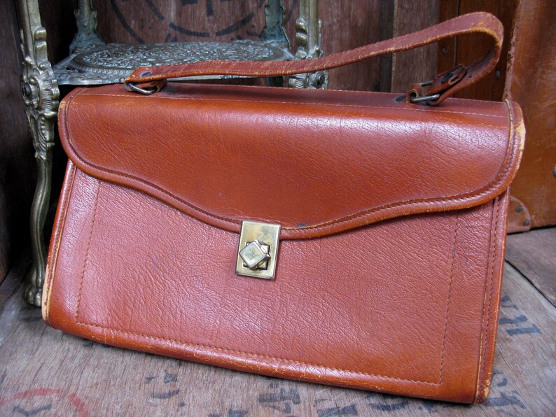 May include: A brown leather satchel with a brass clasp. The bag has a flap closure and a top handle.
