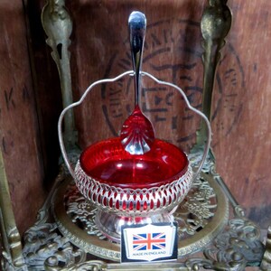 May include: A silver sugar bowl with a red glass liner sits on a decorative metal stand. The stand has a round top and three legs. The sugar bowl has a handle on each side and a spoon inside. The stand has a label that reads "Made in England".