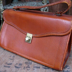 May include: A brown leather satchel with a brass clasp and a strap. The bag has a flap closure and a top handle.