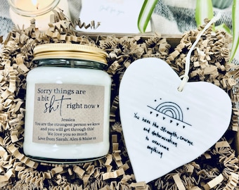 May include: A gift box filled with brown crinkle paper, a white heart-shaped ornament with a rainbow and the text "You have the Strength, courage and determination to overcome anything", and a glass candle jar with a gold lid and a label that reads "Sorry things are a bit shit right now" with the name Jessica and a message of support.