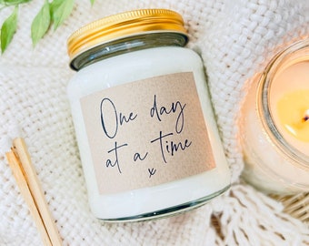 May include: A white candle with a gold lid and a brown label that says "One day at a time" in black script. The candle is sitting on a white fuzzy surface.