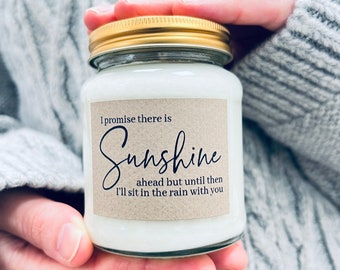 May include: A white candle in a clear glass jar with a gold lid. The jar has a brown paper label with the text "I promise there is Sunshine ahead but until then I'll sit in the rain with you."