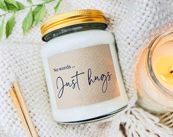 May include: A glass candle jar with a brown label that reads "No words... Just hugs". The candle is white and the jar has a gold lid. The candle is sitting on a white fuzzy surface.