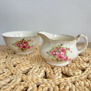 May include: A cream-coloured ceramic sugar bowl and creamer set featuring a delicate floral pattern of pink blossoms and green leaves. Both pieces have a scalloped rim with a thin gold-tone metallic trim along the edges and a glossy finish.