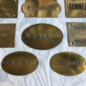 May include: A collection of vintage brass nameplates with engraved text. The plates vary in shape and size, with rectangular and oval designs. Some plates include text such as "L. DENEFFLE CHARPENTIER" and "SONNEZ S.V.P."