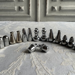 Puede incluir: Una colección de tapones de botella decorativos de metal en varias formas, incluyendo diseños de bellota, cónicos y cilíndricos. Los tapones son plateados y bronceados, dispuestos sobre una superficie blanca estampada. La imagen muestra una variedad de estilos para botellas de vino o bebidas.