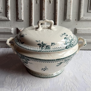 May include: Cream-colored ceramic serving dish with a lid, featuring a floral pattern in teal. The dish has two handles on the sides and one on the lid. The rim and base are decorated with a teal border. A classic piece for serving food.