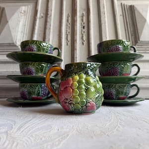 May include: A set of green ceramic teacups and saucers with a grape design, along with a matching creamer pitcher. The teacups and saucers are stacked, and the creamer pitcher is in the foreground. The set is on a white tablecloth.