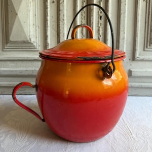 Op de afbeelding: Een ronde pot van geëmailleerd metaal met een levendig kleurverloop dat overgaat van een heldergele bovenkant naar een dieprode basis. De pot is voorzien van een bijpassend deksel met een kleine knop, een zijhandvat en een metalen hengsel.