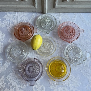 Puede incluir: Ocho exprimidores de limón vintage de vidrio en varios colores, incluyendo transparente, rosa y morado. Los exprimidores tienen un diseño acanalado y están dispuestos sobre un mantel blanco con un patrón floral.
