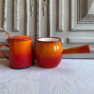 May include: A cylindrical enamel pot with a lid and a side handle, paired with a round enamel saucepan featuring a long handle. Both vessels display a vibrant gradient finish transitioning from a deep red base to a bright orange top.