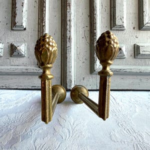 May include: Two antique brass curtain tiebacks. Each tieback has a pinecone-shaped finial. The tiebacks are mounted on a rectangular stem with a curved arm. The background is a white door.