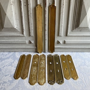 May include: A selection of antique brass door furniture, including long, rectangular plates and smaller keyhole covers. The hardware has a warm, aged patina, with decorative details and keyhole cutouts. The background features a distressed white door and floral fabric.