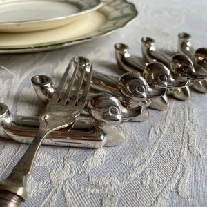 May include: Silverware duck-shaped fork rests. The rests are arranged in a row, with a fork placed on one. The duck rests have a detailed design, and the fork has a decorative handle. A white and green plate is in the background.