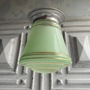Könnte beinhalten: Vintage grüne Glas-Pendelleuchte mit gesprenkelter Oberfläche und goldenen Zierleisten. Die Leuchte hat eine konische Form und einen silberfarbenen Metallsockel. Das Licht hängt an der Decke vor einer weißen Tür.