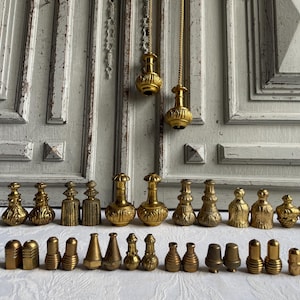 Könnte beinhalten: Eine Sammlung antiker Parfümflaschenverschlüsse und dekorativer Elemente aus Messing. Die Verschlüsse variieren in Form und Design mit aufwendigen Details. Einige sind an Ketten aufgehängt. Die Artikel sind auf einer weißen Oberfläche vor einer Holztür angeordnet.