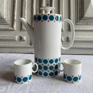 May include: A white ceramic coffee set with a tall coffee pot and two matching mugs. The set features a teal and brown circular pattern around the base and top of the pot and mugs. The coffee pot has a curved handle and a lid.