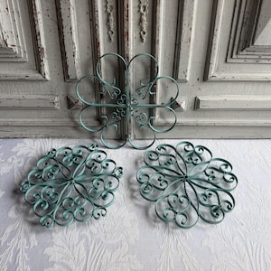 Puede incluir: Tres piezas decorativas de arte mural de metal en color turquesa. La pieza más grande tiene forma de trébol de cuatro hojas. También hay dos piezas redondas más pequeñas con intrincados adornos. Las piezas se exhiben sobre un fondo blanco.