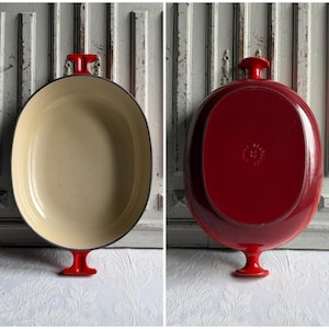 Peut inclure: Un plat à rôtir ovale en fonte émaillée rouge avec un intérieur crème. Le plat a une poignée rouge et une base rouge. Le verso du plat est rouge. Le plat est présenté sur un fond blanc.