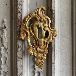 May include: Ornate gold-colored keyhole cover with a floral design. The cover is mounted on a distressed, off-white wooden surface. The keyhole is visible within the decorative frame, suggesting a vintage or antique aesthetic.