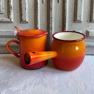 May include: Two vintage enamelware kitchen items. A mug with a lid and a spout, and a small pot with a spout. Both are ombre, transitioning from red to orange. The mug has a handle and the pot has a white interior.