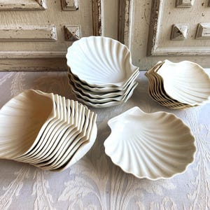 Puede incluir: Una colección de platos blancos con forma de concha. Los platos están apilados en varias pilas, mostrando su diseño único. Las superficies texturizadas y acanaladas de los platos son visibles, lo que sugiere un tema costero o náutico.