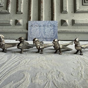 Peut inclure: Un ensemble de cinq repose-couteaux en métal argenté en forme d'oiseaux disposés en rangée horizontale. Chaque pièce présente des ailes détaillées et une tête aviaire distincte, avec une finition métallique polie reflétant la lumière.
