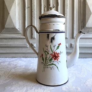 Könnte beinhalten: Eine alte Kaffeekanne aus weißem Emaille mit floralem Design. Die Kanne hat einen langen Ausguss, einen Griff und einen Deckel. Das florale Design umfasst rote Blumen und grüne Blätter. Die Kanne hat ein antikes Aussehen.