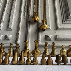 Może przedstawiać: Kolekcja antycznych mosiężnych gałek i uchwytów do drzwi w różnych kształtach. Gałki są eksponowane przed zniszczonymi białymi drzwiami. Dwa pasujące mosiężne uchwyty do drzwi wiszą u góry.