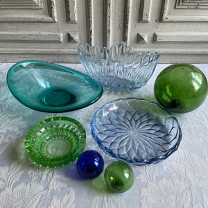 Può includere: Una collezione di ciotole di vetro colorate e sfere decorative. Le ciotole sono in tonalità di blu, verde e verde acqua, con varie forme e trame. Due piccole sfere sono blu e verdi. Gli oggetti sono disposti su una superficie bianca a motivi.