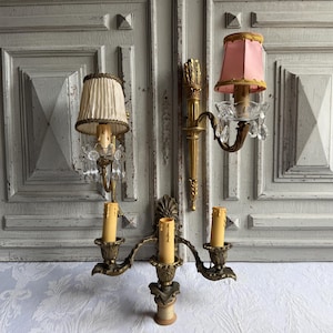 May include: A wall-mounted brass light fixture featuring three candle-style sockets and two fabric lampshades. One shade is cream-colored with pleats, while the other is pink with gold trim. Clear glass crystals hang from the two upper light arms.