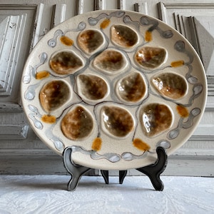 Puede incluir: Un plato para ostras redondo de cerámica con base color crema que presenta doce hendiduras cóncavas en forma de concha esmaltadas en tonos marrón y gris. El borde está decorado con un patrón repetitivo de espirales grises y puntos amarillo mostaza.
