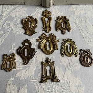 May include: A collection of eight brass keyhole escutcheons featuring various ornate shapes, including oval, rectangular, and shield-like designs with decorative bow and leaf motifs, each with a central opening for a key.