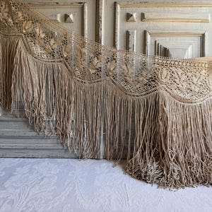 May include: A beige crochet shawl featuring an intricate floral pattern and a long, flowing fringe along the bottom edge. The textile displays a delicate openwork design with scalloped edges, showcasing a neutral tan colour throughout the piece.