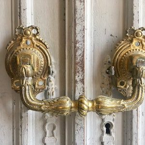 May include: An antique gold-coloured door handle with intricate floral and scroll detailing. The handle is fixed to a distressed, white-painted wooden door. The design includes a curved handle and decorative backplates.
