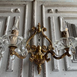 May include: A two-arm wall sconce featuring a polished gold-tone metal frame with ornate scrollwork and a central finial. Two clear glass bobeches hold candle-shaped bulbs, each adorned with dangling faceted crystal prisms and teardrop pendants.